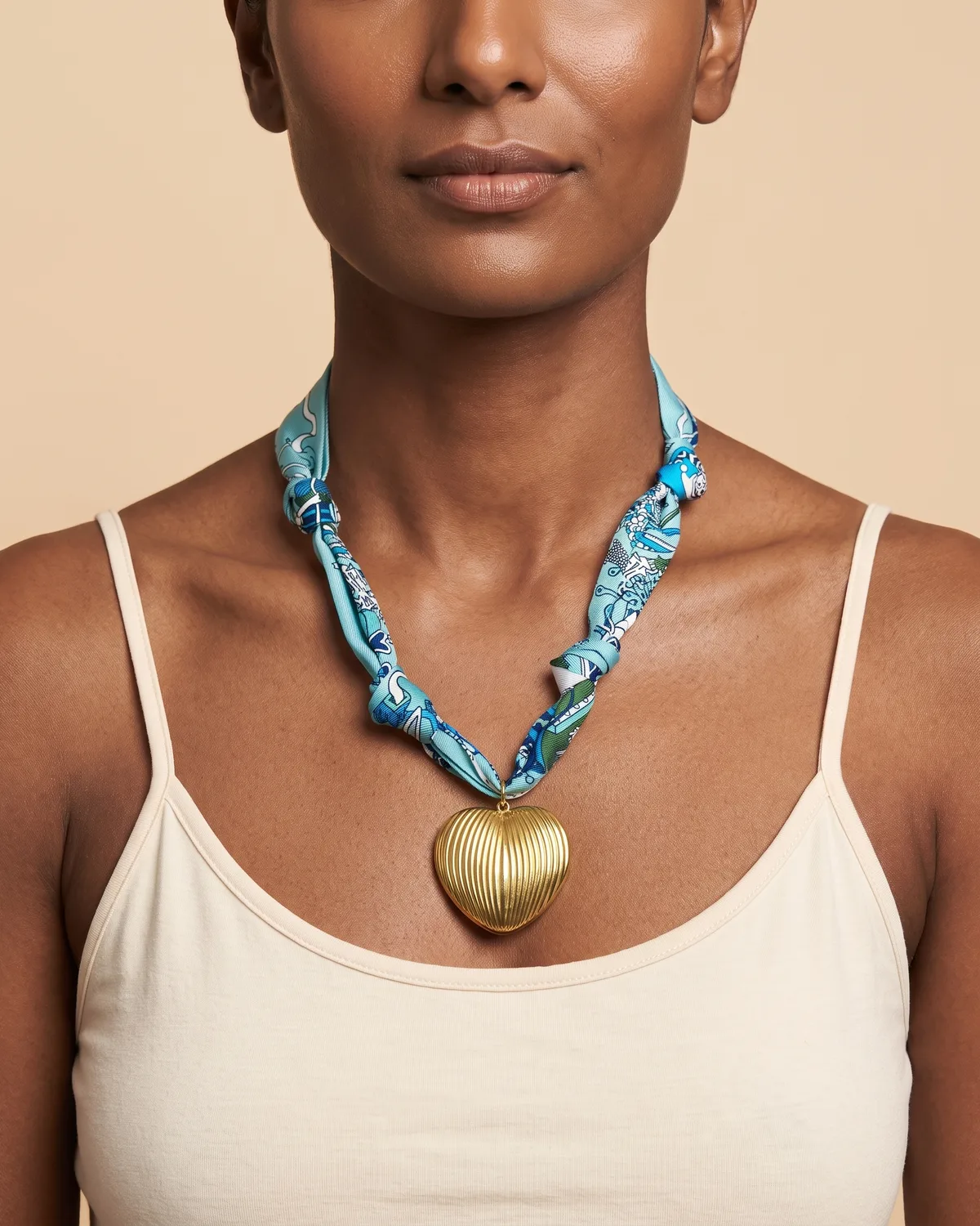Caspian Heart Scarf: A large, ribbed gold heart pendant suspended from a knotted, turquoise-blue fabric ribbon adorned with an intricate white and green graphic pattern.