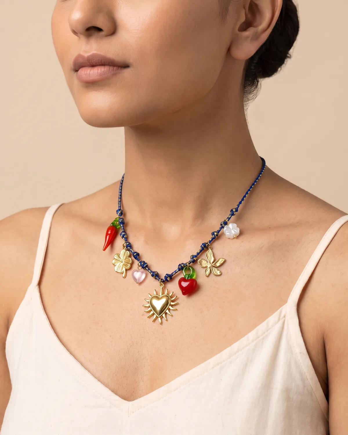 Cherry Blossom Scarf: A slender, dark blue and gold braided cord adorned with an eclectic mix of whimsical pendants, including a gold sun-heart, a four-leaf clover, a red pepper, and a small pink heart.