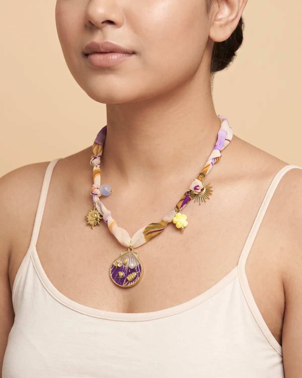 Lavender Lagoon Scarf: a soft cream, purple, and orange patterned fabric ribbon decorated with small gold sun accents and a round gold pendant filled with a purple enamel seashell design.