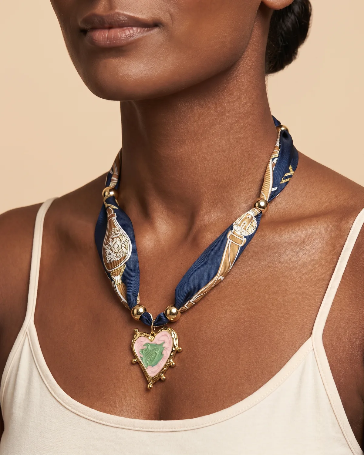Midnight Scarf: a detailed heart-shaped enamel pendant with pink and green accents, suspended from a navy blue and gold patterned fabric scarf-style ribbon interspersed with polished gold beads.