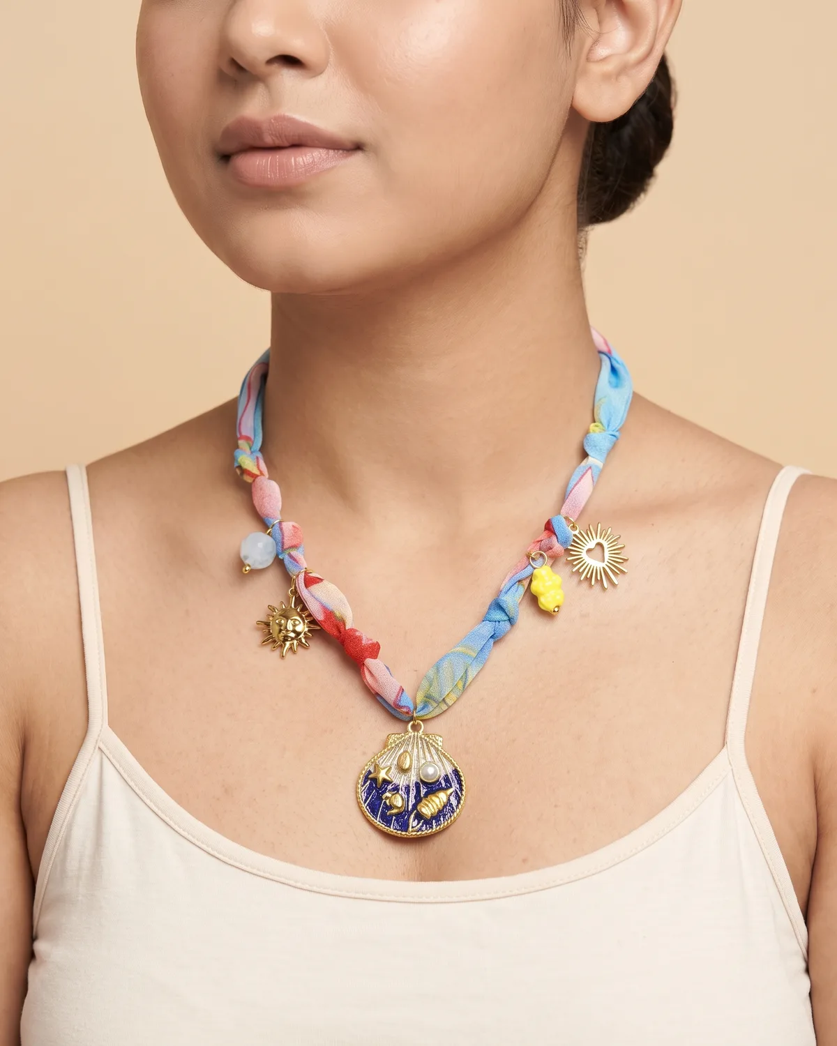 Ocean Wish Scarf: A colorful pastel fabric ribbon studded with small gold sun charms and a prominent, round gold pendant with an intricate blue enamel seashell design.