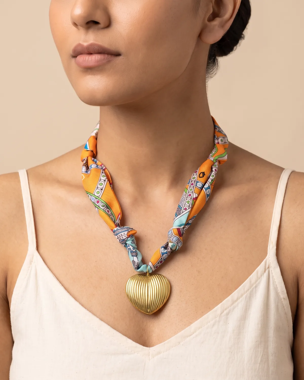 Saffron Love Scarf: a ribbed, gold-toned heart pendant hanging from a knotted orange fabric ribbon adorned with colorful geometric and bohemian patterns.