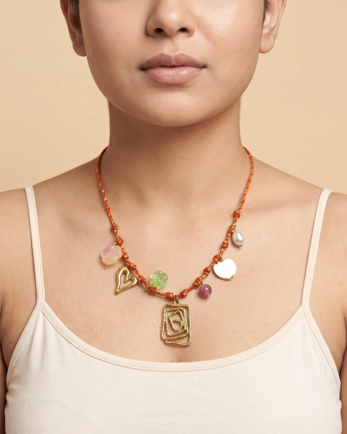 Spiced Honey Charm Scarf: a slender orange and gold braided cord beautifully adorned with an openwork rectangular spiral pendant, a stylized gold heart, and assorted accents including a green flower and a smooth pink bead.