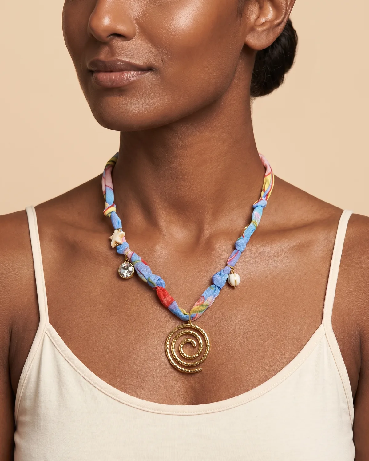 Stardust Spiral Scarf: a gold-toned spiral pendant suspended from a knotted pastel blue and red patterned fabric ribbon that is subtly adorned with small charms, including a round crystal and a tiny starfish.